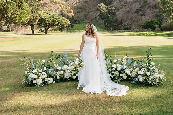 Bride in a white wedding dress standing beside a long floral arrangement of white and blue flowers on a grassy outdoor venue.