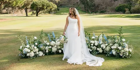 Bride in a white wedding dress standing beside a long floral arrangement of white and blue flowers on a grassy outdoor venue.