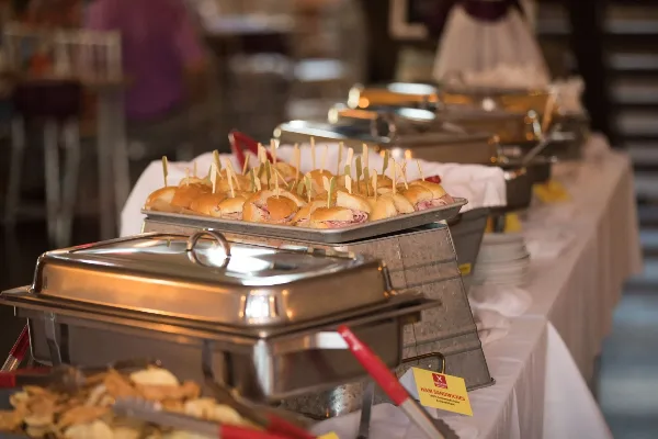 A catering buffet table featuring a tray of ham sandwiches with toothpicks and stainless steel chafing dishes.