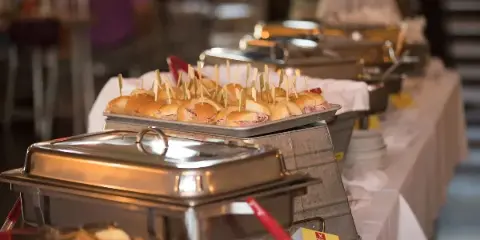 A catering buffet table featuring a tray of ham sandwiches with toothpicks and stainless steel chafing dishes.