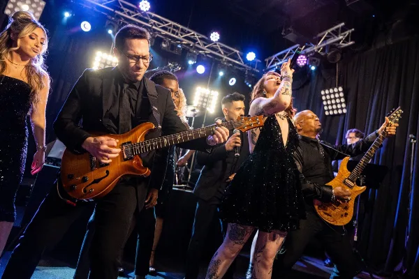 A live band performing on stage with a guitarist, singer, and other musicians under stage lights.