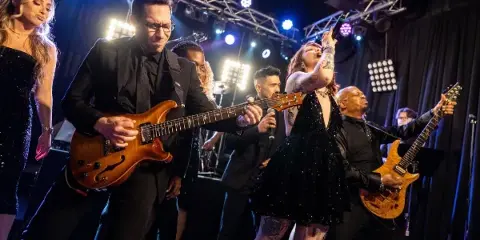 A live band performing on stage with a guitarist, singer, and other musicians under stage lights.