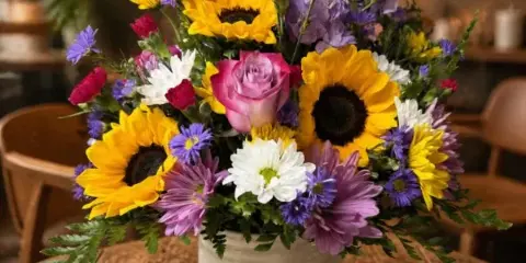A colorful floral arrangement featuring sunflowers, pink roses, and daisies in a white ceramic jar.