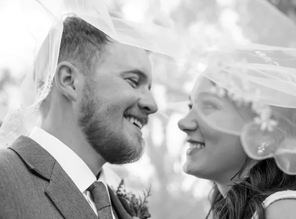 A black and white photograph of a smiling bride and groom looking at each other under a veil.