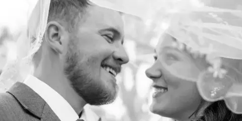 A black and white photograph of a smiling bride and groom looking at each other under a veil.