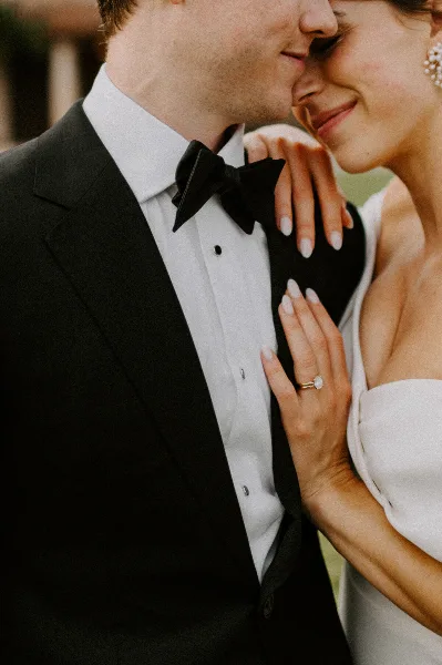 Close-up of a groom in a black tuxedo and a bride in a white dress sharing an intimate moment, with her hand showing an engagement ring.