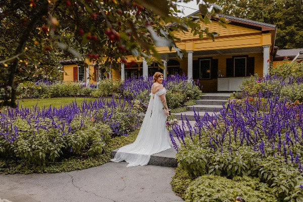 A bride in a white wedding dress standing on a stone path in a garden with purple flowers and a yellow building in the background.