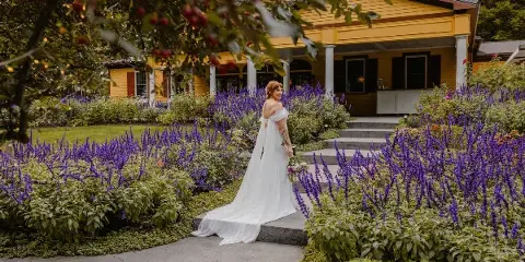 A bride in a white wedding dress standing on a stone path in a garden with purple flowers and a yellow building in the background.
