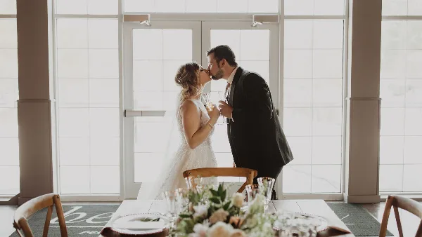 A bride and groom sharing a kiss in an indoor venue with large glass doors and a floral table setting.