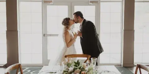 A bride and groom sharing a kiss in an indoor venue with large glass doors and a floral table setting.