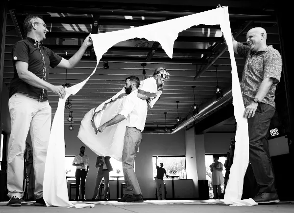 A black and white photograph of a bride and groom laughing and interacting with a large piece of white fabric held by two other men in a modern indoor venue.