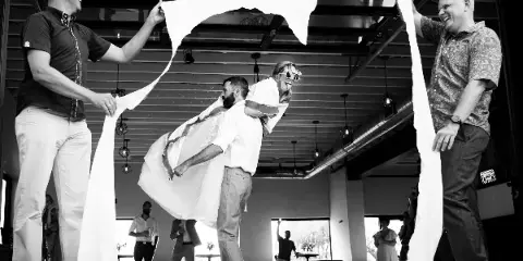 A black and white photograph of a bride and groom laughing and interacting with a large piece of white fabric held by two other men in a modern indoor venue.