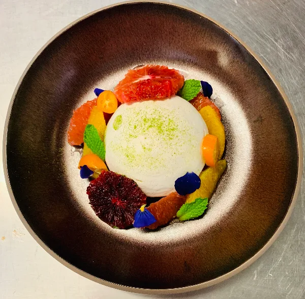 A plated dessert featuring a white mousse or cream center surrounded by citrus segments, mint leaves, and edible flowers on a dark ceramic plate.
