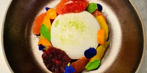 A plated dessert featuring a white mousse or cream center surrounded by citrus segments, mint leaves, and edible flowers on a dark ceramic plate.