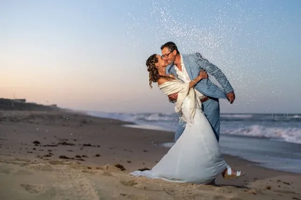 A bride and groom sharing a kiss on a sandy beach with ocean waves in the background.