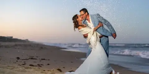 A bride and groom sharing a kiss on a sandy beach with ocean waves in the background.