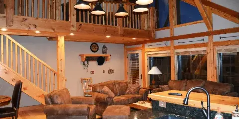 Interior view of a rustic lodge featuring exposed wooden beams, a loft area, leather seating, and a kitchen island.
