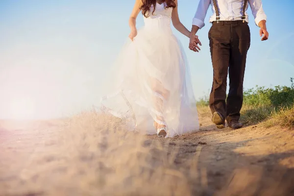 A bride in a white gown and a groom in brown trousers walking hand-in-hand on a sandy path.