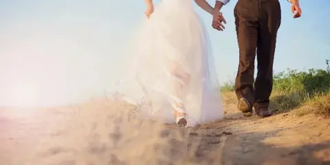 A bride in a white gown and a groom in brown trousers walking hand-in-hand on a sandy path.