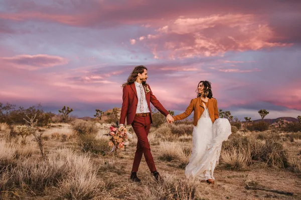 A bride and groom holding hands and walking through a desert landscape at sunset.