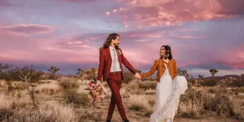 A bride and groom holding hands and walking through a desert landscape at sunset.