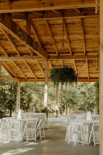 Wedding reception setup with white tables and chairs under a wooden pavilion ceiling with hanging greenery