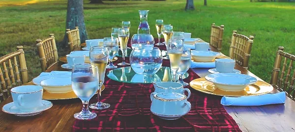 An outdoor event table set with bamboo chairs, blue and gold tableware, and glassware on a grassy lawn.