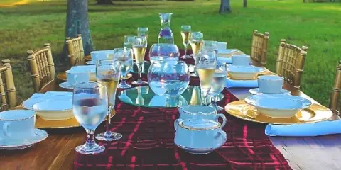 An outdoor event table set with bamboo chairs, blue and gold tableware, and glassware on a grassy lawn.