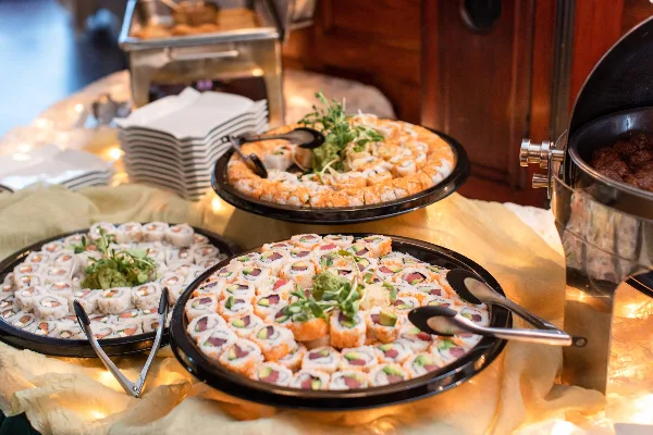 A catering buffet table featuring platters of sushi rolls and other appetizers with serving tongs.