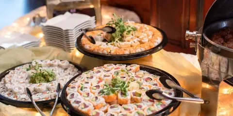 A catering buffet table featuring platters of sushi rolls and other appetizers with serving tongs.