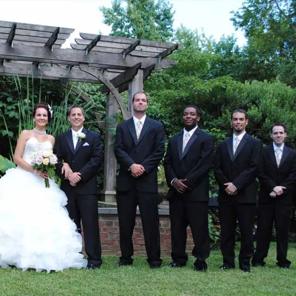 A wedding party consisting of a bride in a white gown and groomsmen in black suits standing outdoors under a wooden pergola.