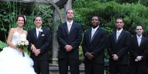 A wedding party consisting of a bride in a white gown and groomsmen in black suits standing outdoors under a wooden pergola.