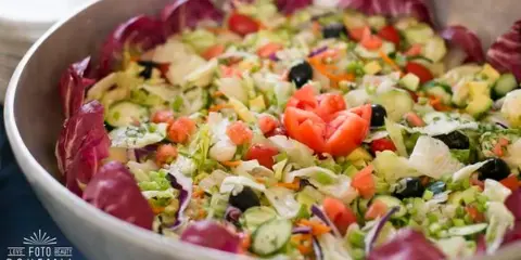 A large catering bowl filled with a fresh salad containing lettuce, tomatoes, cucumbers, and olives.