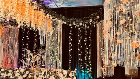 A decorated event backdrop featuring hanging floral garlands, crystal strands, and a floral floor arrangement.