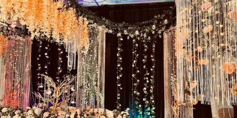 A decorated event backdrop featuring hanging floral garlands, crystal strands, and a floral floor arrangement.