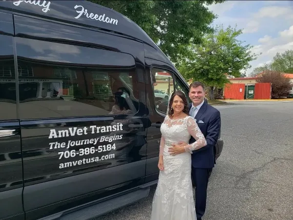A bride and groom posing next to a black AmVet Transit van in a parking lot.