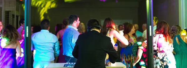 A DJ wearing headphones stands at a mixing console, facing a crowd of wedding guests dancing under colorful event lighting.