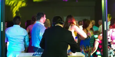 A DJ wearing headphones stands at a mixing console, facing a crowd of wedding guests dancing under colorful event lighting.