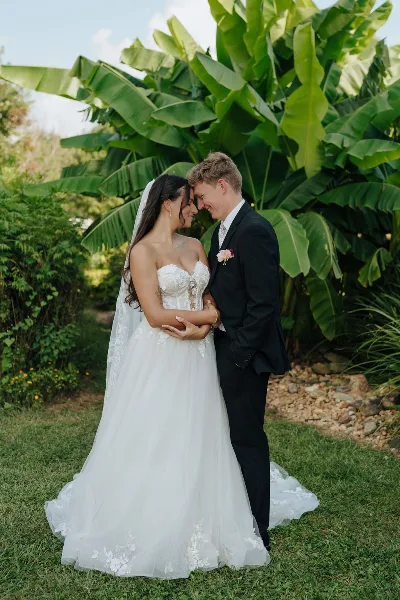 A bride and groom standing close together in a garden setting, with the bride in a white lace gown and the groom in a black suit.