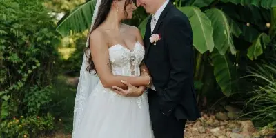 A bride and groom standing close together in a garden setting, with the bride in a white lace gown and the groom in a black suit.