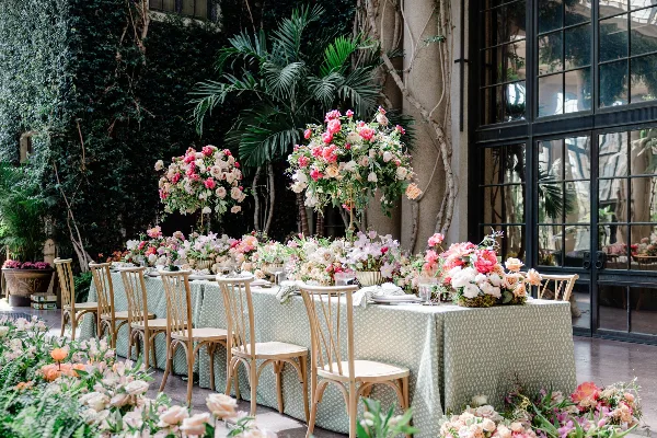 A long banquet table set with a patterned tablecloth, surrounded by wooden chairs and decorated with large, lush floral centerpieces featuring pink and white roses and greenery.