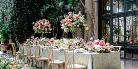 A long banquet table set with a patterned tablecloth, surrounded by wooden chairs and decorated with large, lush floral centerpieces featuring pink and white roses and greenery.