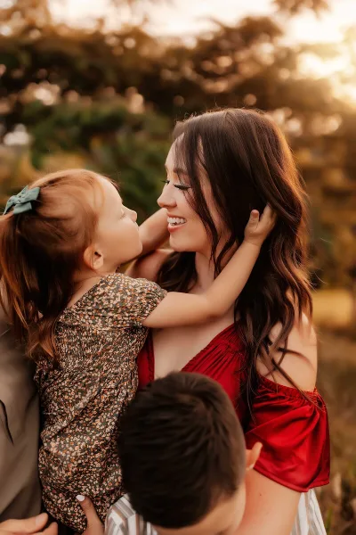 A smiling mother in a red top holding a young child with red hair in a field during golden hour.