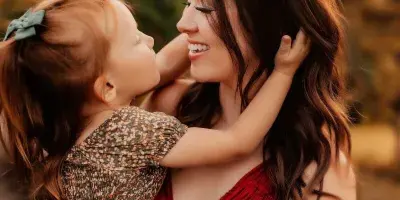 A smiling mother in a red top holding a young child with red hair in a field during golden hour.