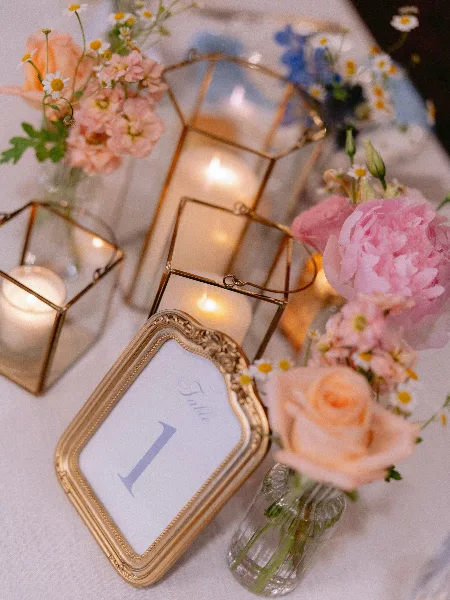 A styled event table featuring lit candles in gold geometric lanterns, a framed 'Table 1' sign, and pink and peach floral arrangements with daisies.