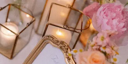 A styled event table featuring lit candles in gold geometric lanterns, a framed 'Table 1' sign, and pink and peach floral arrangements with daisies.