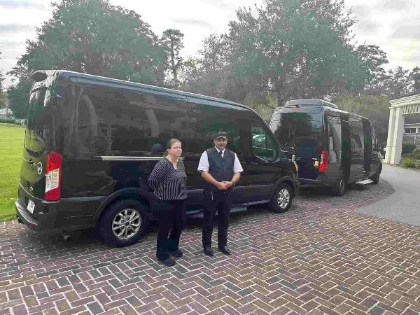 A professional chauffeur in a vest and cap standing next to a black luxury van on a brick driveway.