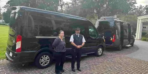A professional chauffeur in a vest and cap standing next to a black luxury van on a brick driveway.