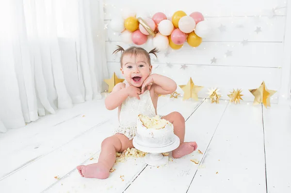 A smiling toddler sitting on a white floor with a small white cake, surrounded by gold stars and pastel balloons.