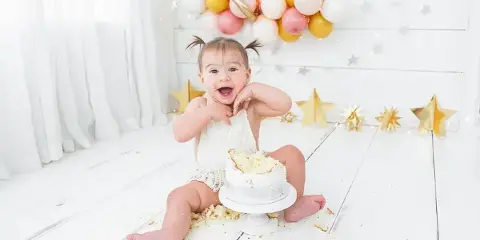 A smiling toddler sitting on a white floor with a small white cake, surrounded by gold stars and pastel balloons.